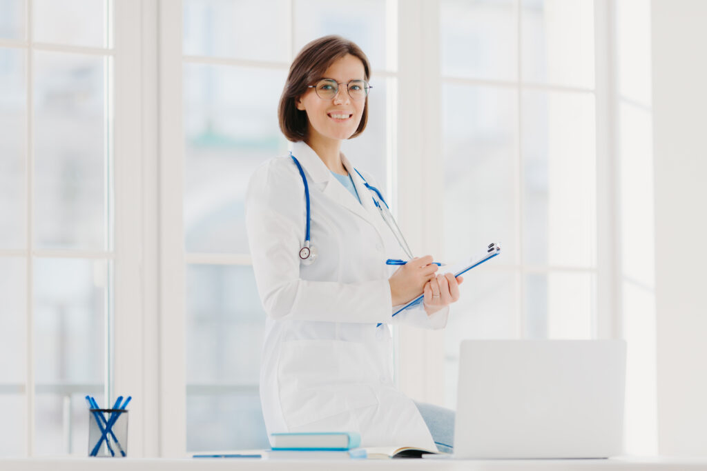 Indoor shot of professional female doctor supplies medic care assistance, poses at desktop with laptop, prepares document agreement paper offer, wears optical glasses and white medical gown.